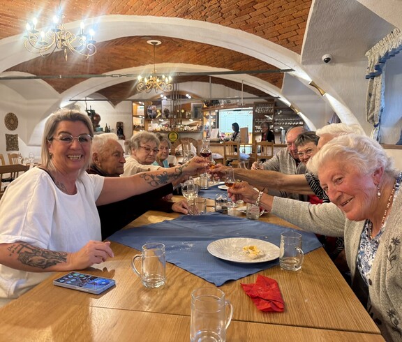 Mehrere Bewohnerinnen und Bewohner sitzen gemeinsam an einem langen Holztisch in einem gemütlichen Gastraum mit Gewölbedecke und stoßen mit kleinen Gläsern an, während vor ihnen leere Gläser, Teller und eine Tischdecke liegen.