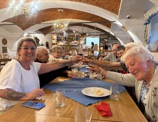 Mehrere Bewohnerinnen und Bewohner sitzen gemeinsam an einem langen Holztisch in einem gemütlichen Gastraum mit Gewölbedecke und stoßen mit kleinen Gläsern an, während vor ihnen leere Gläser, Teller und eine Tischdecke liegen.