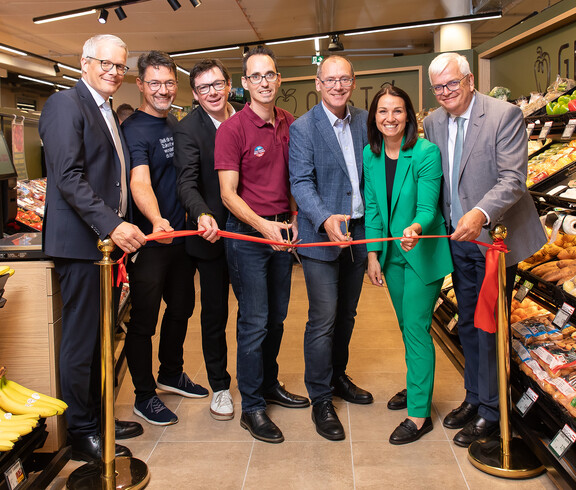 Mehrere Personen schneiden ein rotes Band im Supermarkt zwischen Obstregalen zur Eröffnung durch.