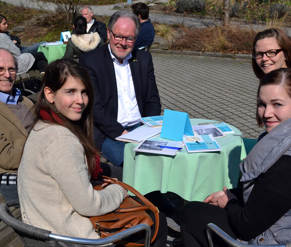 Vier Personen unterschliedlichen Alters und unterschliedlicher Generationen sizen gemütlich im Freien an einem kleinen runden Tisch. 