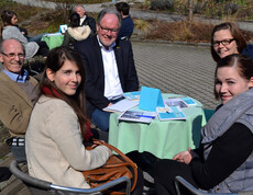 Vier Personen unterschliedlichen Alters und unterschliedlicher Generationen sizen gemütlich im Freien an einem kleinen runden Tisch. 