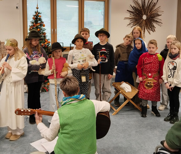 Kindergruppe in Kostümen spielt Adventsszene, Person mit Gitarre begleitet