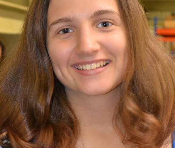 Eine Jugendliche blickt in die Kamera. Sie hat lange braune Haare und trägt ein gestreiftes Oberteil. Sie wirkt fröhlich und lächelt. 