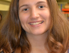 Eine Jugendliche blickt in die Kamera. Sie hat lange braune Haare und trägt ein gestreiftes Oberteil. Sie wirkt fröhlich und lächelt. 