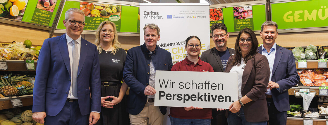 Gruppe in Supermarkt hält Schild „Wir schaffen Perspektiven“ für Caritas-Qualifizierungsprojekt „perspektiven.werk“.