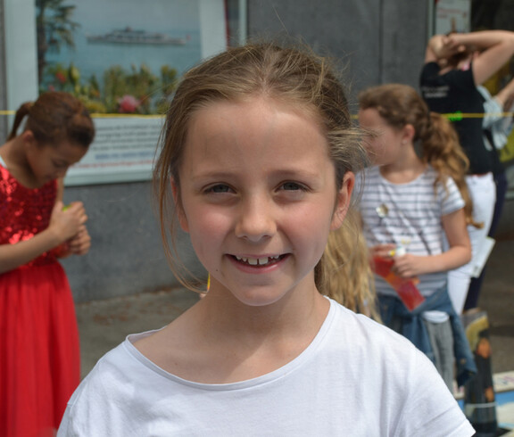 Ein Mädchen im Volksschulalter blickt in die Kamera. Sie hat lange blonde Haare, die sie zurückgebunden hat. Sie trägt ein weißes Oberteil. Im Hintergrund sind drei weitere Mädchen. 