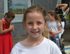 Ein Mädchen im Volksschulalter blickt in die Kamera. Sie hat lange blonde Haare, die sie zurückgebunden hat. Sie trägt ein weißes Oberteil. Im Hintergrund sind drei weitere Mädchen. 