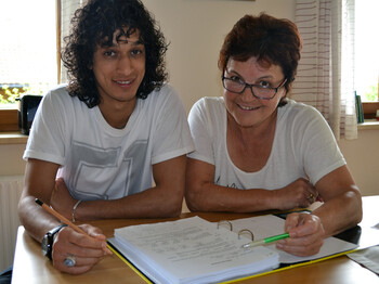 Eine Freiwiliige mittleren Alters hilft einem jungen erwachsenen Flüchtling am Deutschlernen. Sie sitzen entspannt an einem Holztisch, auf dem sich ein großer Ordner voller Lernmaterialien, befindet. Der Flüchtling hat lange schwarze, die Freiwillige kurze rot gefärbte Haare. Beide tragen weiße Oberteile. 