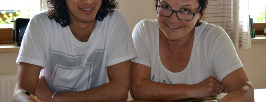 Eine Freiwiliige mittleren Alters hilft einem jungen erwachsenen Flüchtling am Deutschlernen. Sie sitzen entspannt an einem Holztisch, auf dem sich ein großer Ordner voller Lernmaterialien, befindet. Der Flüchtling hat lange schwarze, die Freiwillige kurze rot gefärbte Haare. Beide tragen weiße Oberteile. 