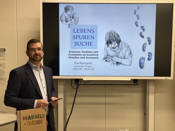 Mann mit dunklen kurzen Haaren im Anzug mit weißem Hemd seitlich links von einem Präsentationsbildschirm.