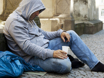 Ein Mann mittleren Alters sitzt auf Pappe in einer gepflasterten Straße. Er hat einen grauen Hoodie an. Die Kapuze trägt er auf dem Kopf, der zum Boden geneigt ist. In seiner rechten Hand hält er einen weißen Plastikbecher. Seine Kleidung ist ein wenig verschmutzt. Dem Anschein nach ist er ein Bettler. 