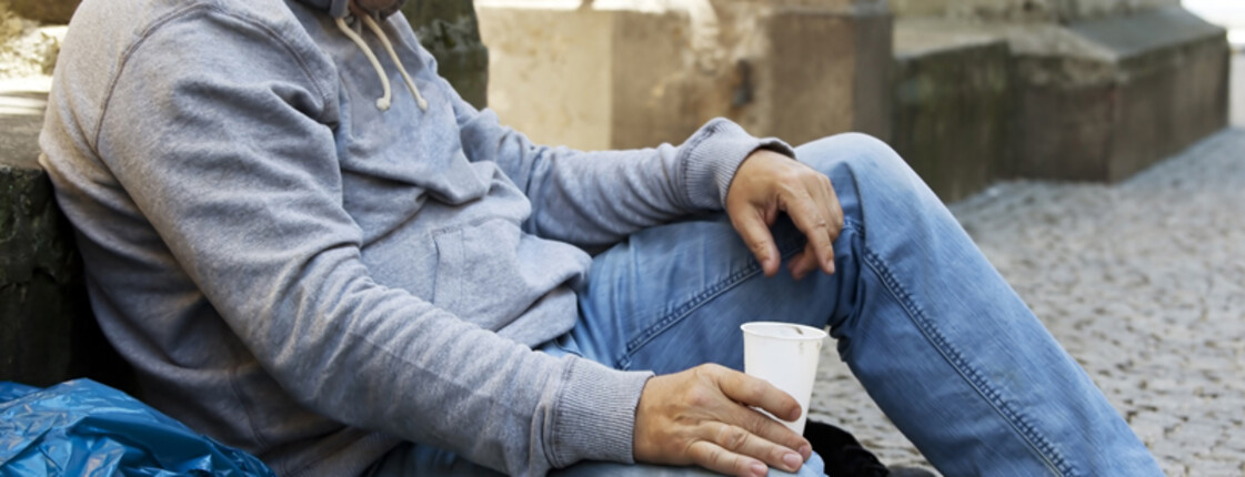 Ein Mann mittleren Alters sitzt auf Pappe in einer gepflasterten Straße. Er hat einen grauen Hoodie an. Die Kapuze trägt er auf dem Kopf, der zum Boden geneigt ist. In seiner rechten Hand hält er einen weißen Plastikbecher. Seine Kleidung ist ein wenig verschmutzt. Dem Anschein nach ist er ein Bettler. 