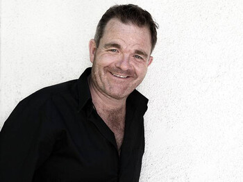 Ein Herr mittleren Alters lacht in die Kamera. Er hat kurze Haare und trägt ein schwarzes Hemd.