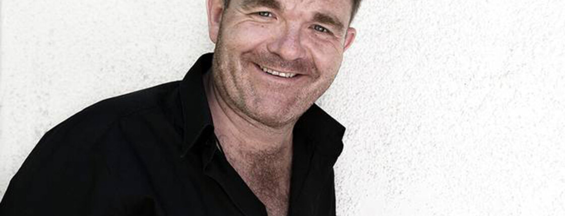 Ein Herr mittleren Alters lacht in die Kamera. Er hat kurze Haare und trägt ein schwarzes Hemd.