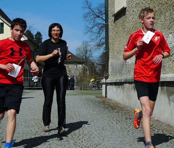 Drei Personen laufen in Sportbekleidung beim Stundenlauf. Eine Frau in der Mitte ganz schwarz bekleidet und zwei Jungs rennen ihr davon.
