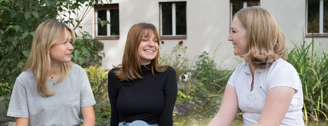 Drei junge Frauen sitzen in einem Garten auf einer niedrigen Steinmauer und unterhalten sich.