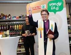 Geistlicher segnet mit Weihwasser den Markt, steht mit Buch vor SPAR-Wand und Microfonständer.
