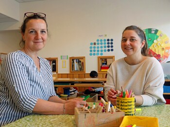 Zwei Frauen sitzen an einem Tisch in einem Kindergarten und lächeln in die Kamera.