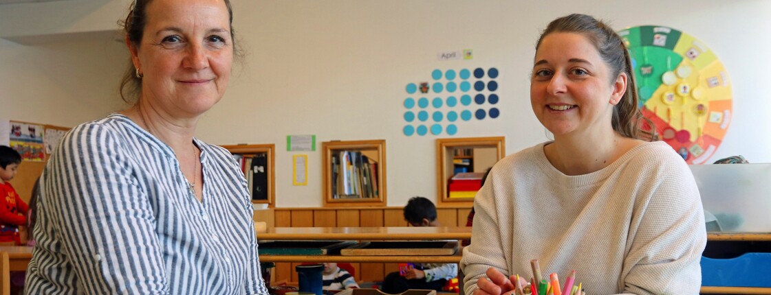 Zwei Frauen sitzen an einem Tisch in einem Kindergarten und lächeln in die Kamera.