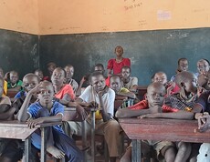 Eine Gruppe südsudanesischer Burschen sitzt in einem Schulzimmer und blickt zur Kamera. Hinten steht eine Frau in einem roten Kleid. 