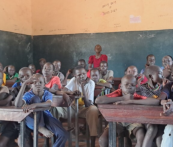 Eine Gruppe südsudanesischer Burschen sitzt in einem Schulzimmer und blickt zur Kamera. Hinten steht eine Frau in einem roten Kleid. 