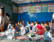 Zahlreiche Kinder sitzen auf dem Boden eines Klassenzimmers mit bunten Lernpostern an der Wand, während eine Lehrerin an der Tafel steht.