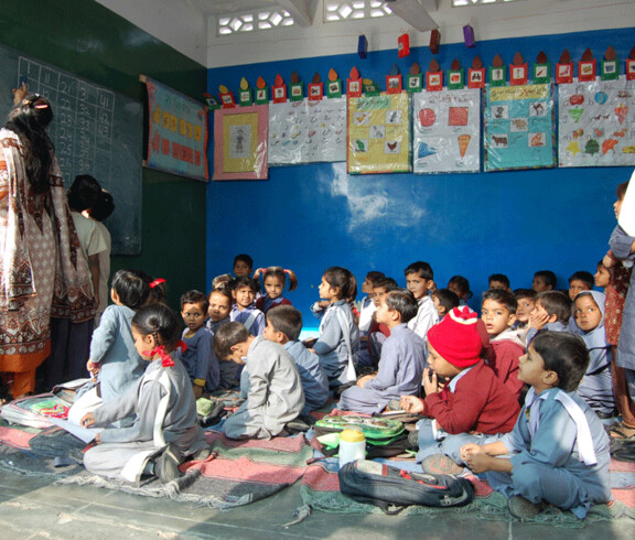 Zahlreiche Kinder sitzen auf dem Boden eines Klassenzimmers mit bunten Lernpostern an der Wand, während eine Lehrerin an der Tafel steht.