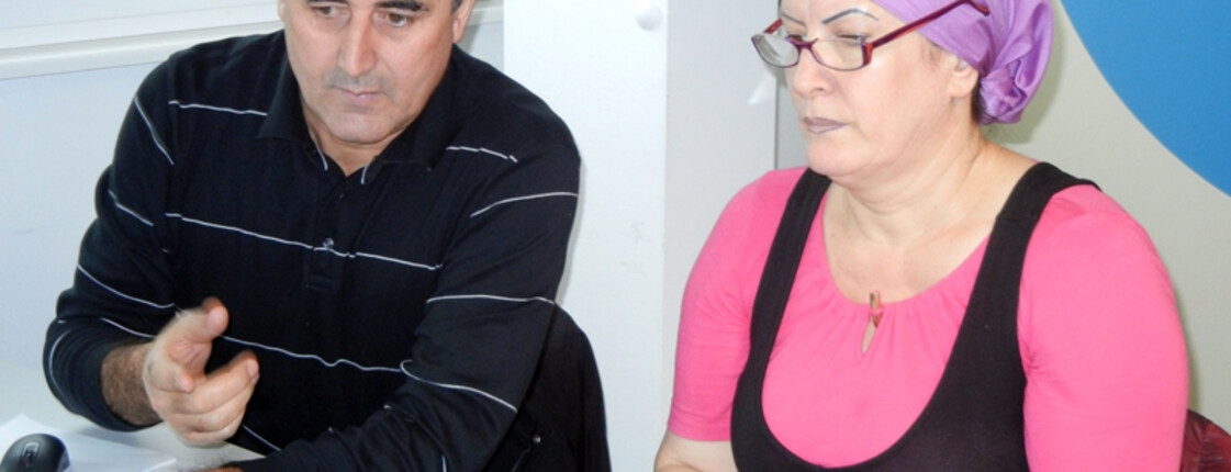 Zwei Flüchtlinge, ein Mann und eine Frau, sitzen an einem Tisch und sind sich am Unterhalten. Der Mann trägt einen schwarzen Pullover, die Frau ein schwarz-rosa Oberteil mit einem violetten Kopftuch. 