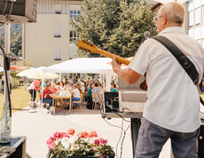 Musiker spielt Gitarre auf Bühne im Freien, Publikum sitzt an Tischen unter Zelten beim Sommerfest