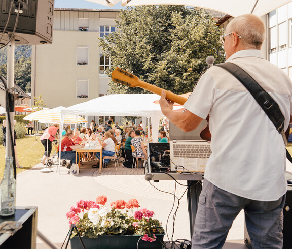 Musiker spielt Gitarre auf Bühne im Freien, Publikum sitzt an Tischen unter Zelten beim Sommerfest