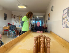 Bewohner im Rollstuhl hält eine Kugel, während Kegel am Ende der langen Holzbahn stehen.
