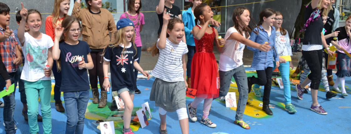 Bregenzer Bürgermeister Markus Lienhart steht mit Kindern im Volksschulalter im Freien. Sie schauen sich ein großes Kunststück an, das auf Asphalt ausgelegt worden ist. Es repräsentiert die Weltkugel und ist somit blau grün und braun. Auf den jeweiligen Kontinenten sind Kärtchen aufgestellt. Auf diesen sind Kinder zu sehen. 