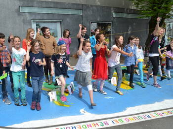 Bregenzer Bürgermeister Markus Lienhart steht mit Kindern im Volksschulalter im Freien. Sie schauen sich ein großes Kunststück an, das auf Asphalt ausgelegt worden ist. Es repräsentiert die Weltkugel und ist somit blau grün und braun. Auf den jeweiligen Kontinenten sind Kärtchen aufgestellt. Auf diesen sind Kinder zu sehen. 