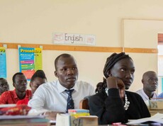 Mehrere Männer und Frauen sitzen aufmerksam in einem Klassenraum mit englischsprachigen Lernpostern an der Wand und folgen konzentriert dem Unterricht im Lehrerausbildungszentrum in Yambio.