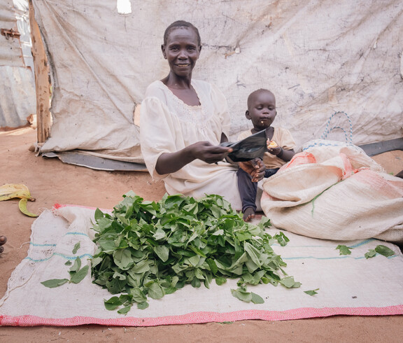 Eine Frau im Südsudan sitzt am Boden auf einem weißen Tuch und hält ein Kleinkind am Arm. Vor ihr liegen auf einem Tuch viele grüne Blätter.