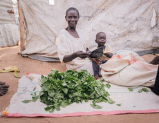 Eine Frau im Südsudan sitzt am Boden auf einem weißen Tuch und hält ein Kleinkind am Arm. Vor ihr liegen auf einem Tuch viele grüne Blätter.