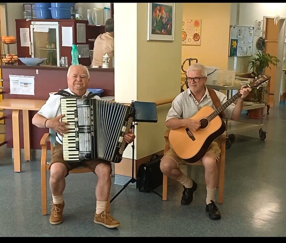 Zwei Personen sitzen in einem Aufenthaltsbereich eines Pflegewohnhauses und sorgen mit Akkordeon und Gitarre für musikalische Unterhaltung.
