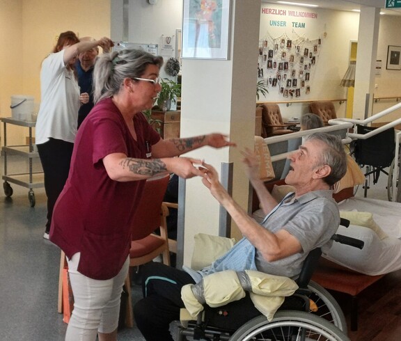 Eine Pflegekraft tanzt mit einem Bewohner im Rollstuhl und sorgt gemeinsam mit weiteren Personen im Hintergrund für eine fröhliche Atmosphäre beim „Tanz in den Mai“ im Pflegewohnhaus.
