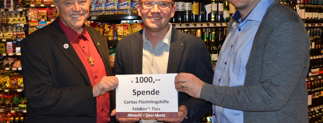 Zwei Männer übergeben in einem SPAR Supermarkt eine Spende von 1000 Euro der Caritas Martin Fellacher, fachbereichsleiter der Flüchtlings- und Migrantenhilfe. Sie stehen vor einem mit Wein beifüllten Regal und lächeln in die Kamera. 