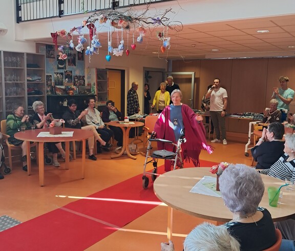 Eine Person präsentiert mit einer Gehhilfe auf einem roten Laufsteg eine Modeschau im barrierefreien Gemeinschaftsraum des Pflegewohnhauses, während das Publikum ringsum an Tischen sitzt.