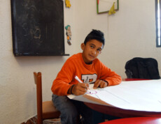 Ein Junge sitzt an einem Tisch im Kinderzentrum Eden, schreibt auf ein Blatt Papier und schaut nachdenklich.