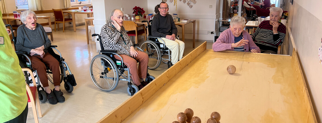 Gruppe von Bewohner*innen verfolgen eine Tischkegelkugel auf der Holzbahn im Aufenthaltsbereich.
