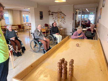 Gruppe von Bewohner*innen verfolgen eine Tischkegelkugel auf der Holzbahn im Aufenthaltsbereich.