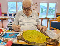 Person bemalt eine große Papiermaché-Schildkröte mit gelber Farbe, daneben liegen Pinsel, Farben und Vorlagen.
