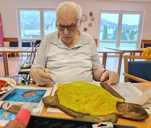 Person bemalt eine große Papiermaché-Schildkröte mit gelber Farbe, daneben liegen Pinsel, Farben und Vorlagen.