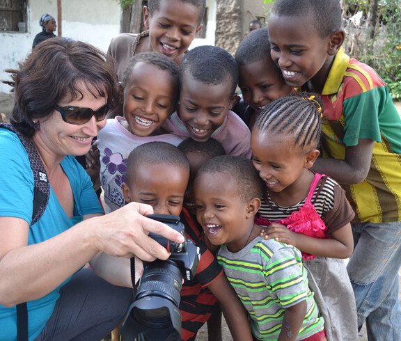 Caritas-Mitarbeiterin Elke Kager zeigt äthiopischen Kindern Fotos auf ihrer Kamera. Die Kinder schauen gespannt und sind erfreut. Elke trägt eine blaue Bluse und die Kinder sind bunt, in unterschiedlichen Farben gekleidet. 