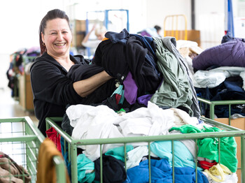 Eine carla Mitarbeiterin mittleren Alters mit langen schwarzen Haaren sortiert fröhlich und heiter Kleidung aus einem grünen Schiebewagen. 