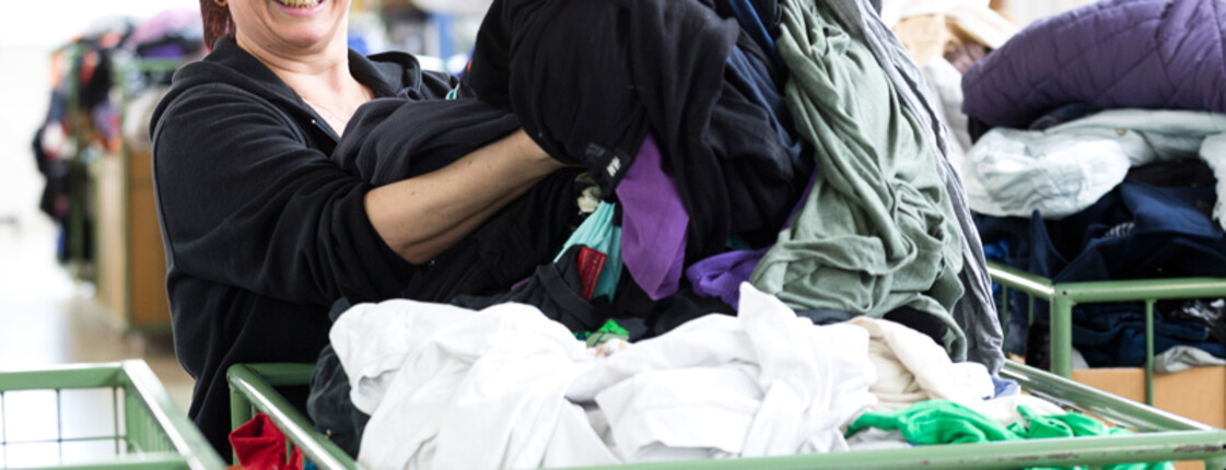 Eine carla Mitarbeiterin mittleren Alters mit langen schwarzen Haaren sortiert fröhlich und heiter Kleidung aus einem grünen Schiebewagen. 