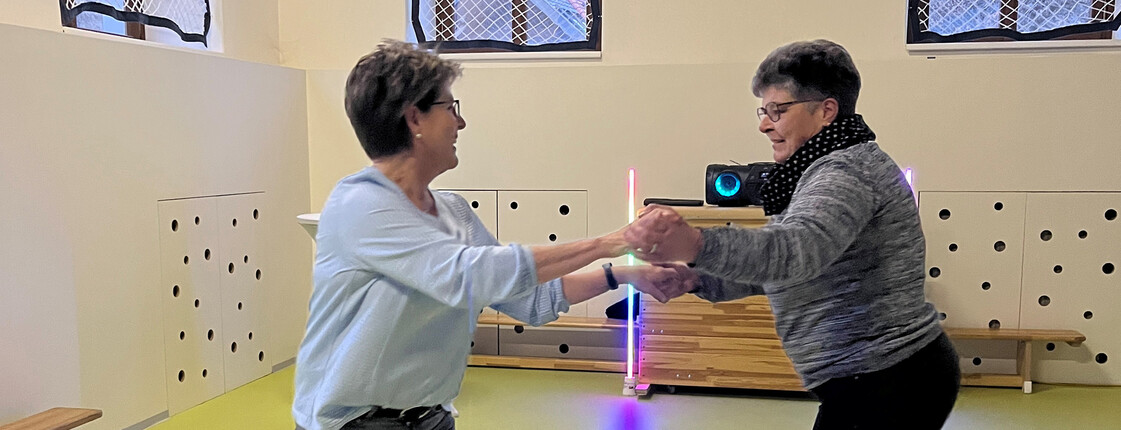 Zwei Damen halten sich an den Händen und tanzen in einem Turnsaal.