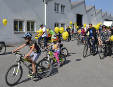 Ca. 20 Menschen mit an ihren Rädern gelb befestigten Luftballonen befinden sich auf einem großen asphaltierten Platz. 